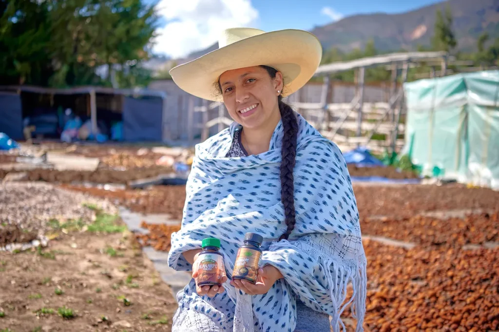 Una mujer agricultora sonriente de Cajamarca de pie en un campo verde, sosteniendo un pu&ntilde;ado de Huanarpo Macho rojo reci&eacute;n cosechado, mostrando nuestra obtenci&oacute;n sostenible.